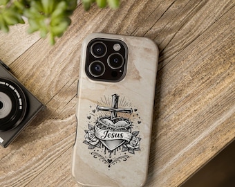 Può includere: Custodia per telefono con un design marmorizzato. Presenta una grafica in bianco e nero di un cuore, una croce e rose, con la parola "Jesus" su uno striscione. La custodia è su una superficie di legno.
