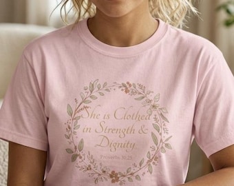 Può includere: T-shirt rosa chiaro con un motivo a ghirlanda floreale. Il testo all'interno della ghirlanda recita "She is Clothed in Strength & Dignity" e "Proverbi 31:25". Il design è nei toni del marrone e del verde.