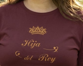 Può includere: T-shirt bordeaux con testo dorato e design a corona. Il testo recita "Hija del Rey" e "SALMO 45:13". La maglietta è a girocollo.