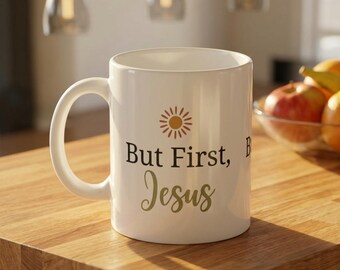 Può includere: Tazza in ceramica bianca con la scritta "But First, Jesus" in nero e oro, insieme a una grafica a forma di sole. La tazza ha un manico grande ed è appoggiata su una superficie di legno.