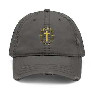 Könnte beinhalten: Verwischte graue Baseballkappe mit dem Text "CHRIST IS KING" in Gelb und einem gelben Kreuzemblem. Die Kappe hat einen getragenen Look mit ausgefransten Kanten.