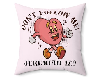 Può includere: Un cuscino quadrato rosa chiaro con il testo "DON'T FOLLOW ME!" e "JEREMIAH 17:9" in nero. Il cuscino presenta un personaggio a forma di cuore dei cartoni animati con braccia, gambe e un gesto di pollice in su. Il cuore è rosa con dettagli gialli.