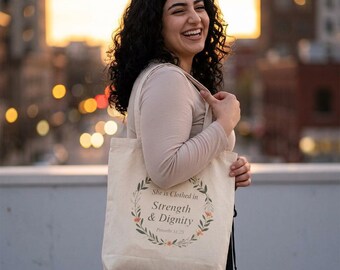 Può includere: Borsa tote in tela beige con un design a ghirlanda floreale e la scritta "She is Clothed in Strength & Dignity Proverbs 31:25". La borsa è tenuta da una persona.