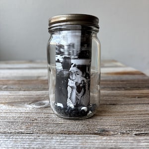 May include: A clear glass jar with a metal lid containing a black and white photograph. The photograph features a person with their hand on their chin. Small black and white objects are at the bottom of the jar.