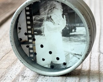 May include: A small, cylindrical, silver-toned container with a black and white photograph visible through a clear window. The photograph depicts a person. The container has a distressed, vintage appearance.