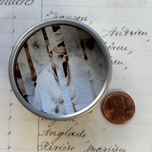 May include: A small, round, silver tin with a sepia-toned image of a person wearing sunglasses and a pearl necklace. A copper penny is next to the tin. The image is partially obscured by vertical strips.