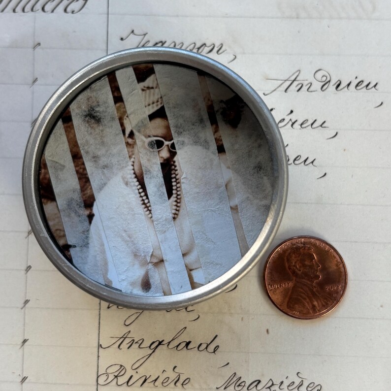 May include: A small, round, silver tin with a sepia-toned image of a person wearing sunglasses and a pearl necklace. A copper penny is next to the tin. The image is partially obscured by vertical strips.