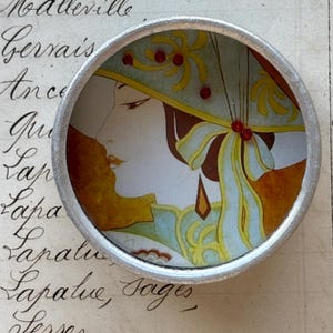 Könnte beinhalten: Ein runder, silberfarbener Kompaktspiegel mit einer Illustration im Art-Déco-Stil einer Frau im Profil. Die Frau trägt einen Hut mit gelben und roten Akzenten. Der Hintergrund ist ein Vintage-Papier mit handschriftlichem Text.