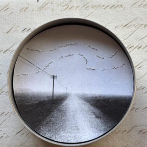 May include: A black and white circular art piece featuring a rural landscape with a road leading to the horizon. A telephone pole and power lines are visible. The artwork is set within a silver metal frame.