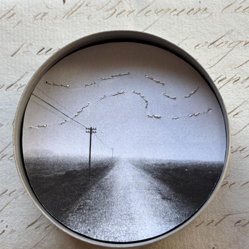 May include: A black and white circular art piece featuring a rural landscape with a road leading to the horizon. A telephone pole and power lines are visible. The artwork is set within a silver metal frame.