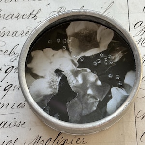 Könnte beinhalten: Ein kleiner, runder, silberfarbener Behälter mit einem Schwarz-Weiß-Bild im Inneren. Das Bild zeigt ein Blumenmuster und eine Silhouette einer Person. Kleine, klare, runde Verzierungen sind über das Bild verstreut.