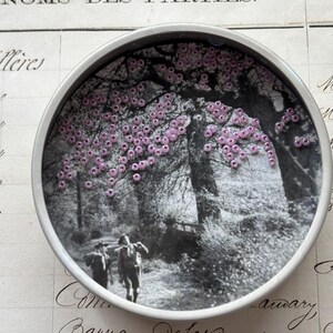 May include: A round, silver-rimmed decorative piece featuring a black and white image of a path through a forest with two figures walking. Pink beads are arranged to resemble blossoms in the tree canopy.