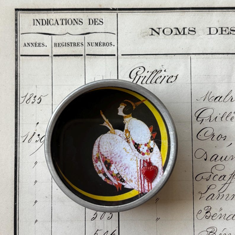 K&ouml;nnte beinhalten: Ein runder Knopf mit silbernem Rand, der eine Vintage-Illustration einer Frau in einem wei&szlig;en Kleid mit rotem Herz auf schwarzem Hintergrund zeigt. Der Knopf befindet sich auf altem Registerpapier.