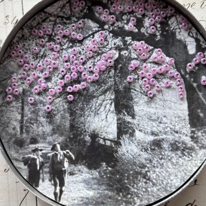 May include: A round, black and white image of a forest scene with two figures walking on a path. The image is embellished with small, pink beads, clustered to resemble blossoms on a tree. The scene is framed by a metal edge.