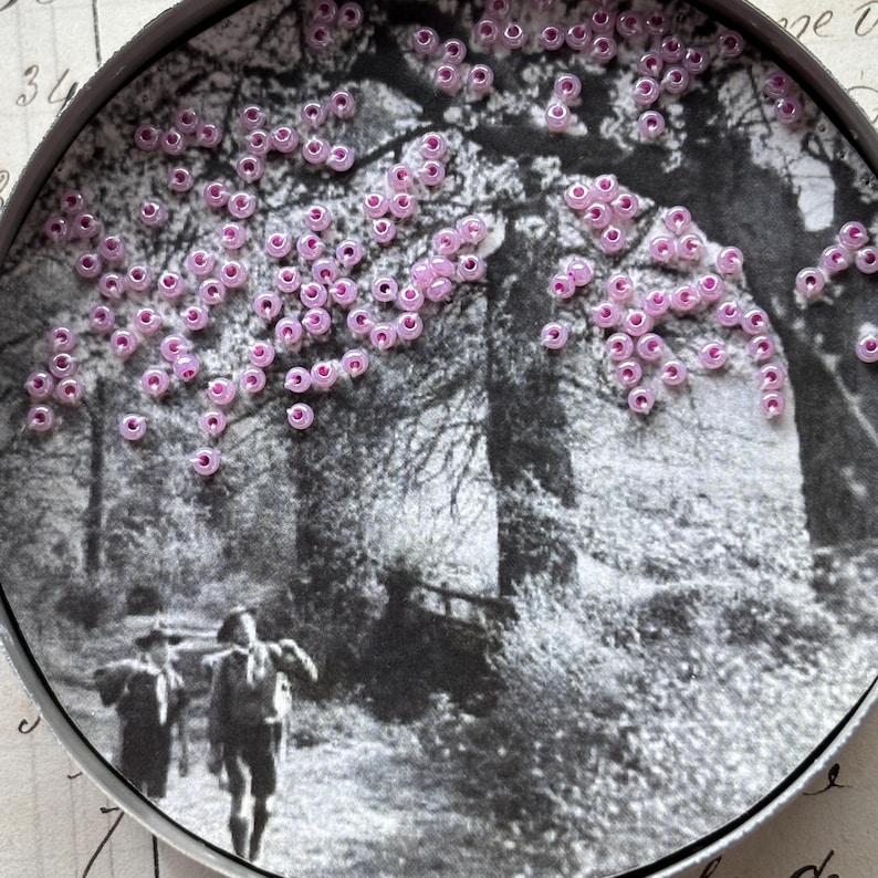 May include: A round, black and white image of a forest scene with two figures walking on a path. The image is embellished with small, pink beads, clustered to resemble blossoms on a tree. The scene is framed by a metal edge.