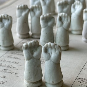 May include: A collection of white porcelain doll arms with clenched fists. The arms are arranged in a row on a piece of paper with handwritten text.