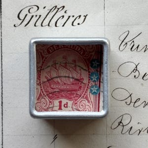 May include: A silver-toned square pendant showcasing a vintage Bermuda stamp. The stamp shows a red sailing ship, with the words "BERMUDA" and "1d". Two blue star-shaped decorations are on the right side of the stamp.