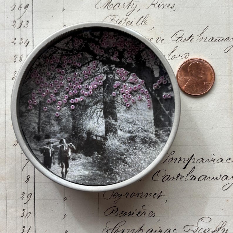 May include: A round, silver-toned metal container with a black and white image of a path through a forest. The image is embellished with small, pink beads. A penny is next to the container.