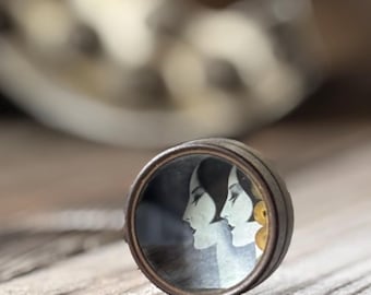 May include: A round, metal locket with a glass window showing a black and white image of two women's faces. The women are facing each other, and their hair is styled in a similar fashion. The locket has a gold-colored rim.