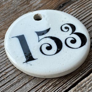 May include: A round, white ceramic house number plaque with the number 153 in black, stylized font. The plaque has a small hole at the top for hanging. The background is a weathered wooden surface.