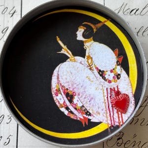 K&ouml;nnte beinhalten: Ein runder Metallbeh&auml;lter mit einer Vintage-Illustration einer Frau in einem wei&szlig;en Kleid, vor einem schwarzen Hintergrund mit einem gelben Halbmond. Ein roter Herzanh&auml;nger h&auml;ngt am Kleid.