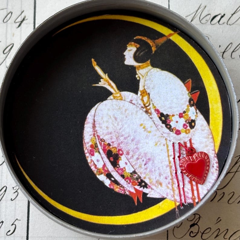 K&ouml;nnte beinhalten: Ein runder Metallbeh&auml;lter mit einer Vintage-Illustration einer Frau in einem wei&szlig;en Kleid, vor einem schwarzen Hintergrund mit einem gelben Halbmond. Ein roter Herzanh&auml;nger h&auml;ngt am Kleid.