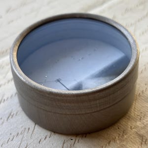 May include: A small, round, silver-colored container with a clear, blue-tinted lid. The container has a brushed metal appearance and a simple, minimalist design. The lid appears to be made of glass or a similar transparent material.