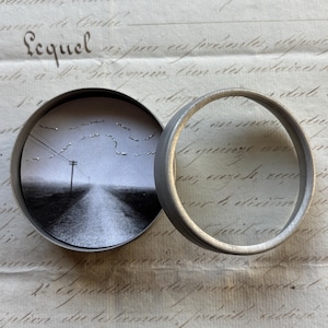 May include: A small, round, silver-toned metal container with a hinged lid, open to reveal a black and white photograph of a road receding into the distance. The lid is open, and the container sits on a vintage paper background.