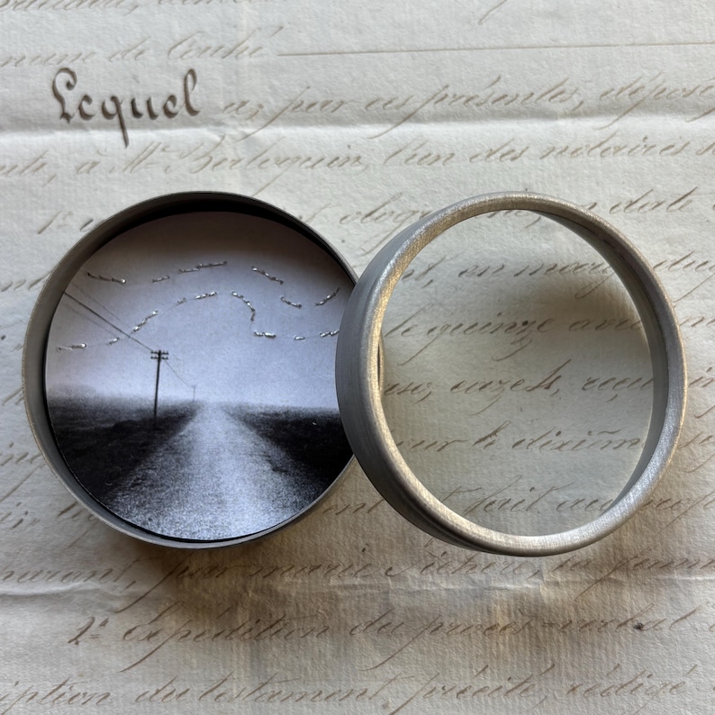 May include: A small, round, silver-toned metal container with a hinged lid, open to reveal a black and white photograph of a road receding into the distance. The lid is open, and the container sits on a vintage paper background.