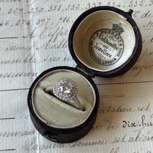 May include: A diamond engagement ring in a black, round, hinged jewelry box. The ring features a large, round, halo-style diamond. The box interior is lined with light green velvet. The box lid has a printed logo: "J.J. DURRANT & SON, Goldsmiths & Jewellers, 126 CHEAPSIDE, LONDON."