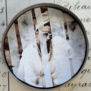 May include: A circular metal container featuring a sepia-toned image of a person wearing sunglasses and a pearl necklace. The image is divided by vertical white strips. The background includes handwritten script.