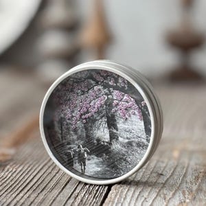 May include: A small, round, silver tin with a black and white image of a person walking on a path under a tree with pink blossoms. The tin is sitting on a wooden surface.