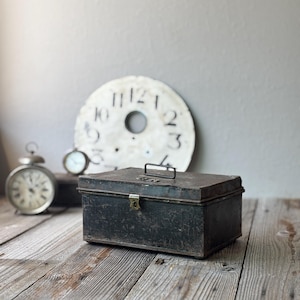 May include: A weathered, dark metal box with a handle and clasp sits on a wooden surface. A large, antique clock face and two smaller clocks are in the background. The scene evokes a vintage aesthetic.