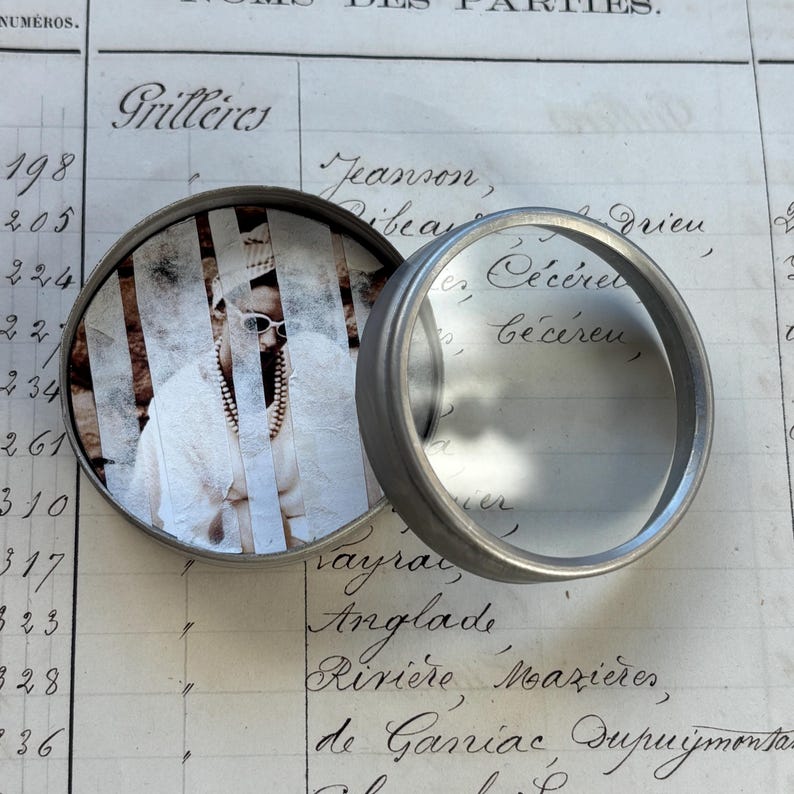 May include: A small, round, silver-toned metal container with a hinged lid, partially open. The lid's interior displays a sepia-toned image of a person wearing sunglasses and a pearl necklace. The container rests on a vintage ledger.