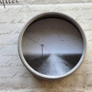 May include: A small, round, silver-colored tin containing a black and white image of a rural road receding into the distance under a cloudy sky. A telephone pole and power lines are visible.