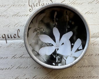 May include: A round, silver-toned metal container with a black and white floral design. The image features white flowers with sparkling embellishments. The container rests on a background of vintage script.