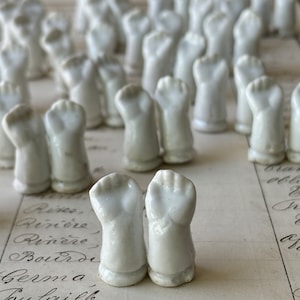 May include: A collection of white porcelain doll arms, each with a closed fist, arranged in rows on a piece of paper with handwritten text.