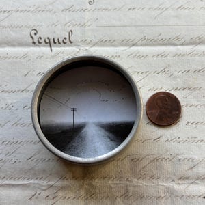 May include: A small, round, silver-colored tin containing a black and white landscape drawing of a road receding into the distance under a cloudy sky. A penny is next to the tin. The drawing features a telephone pole and power lines.