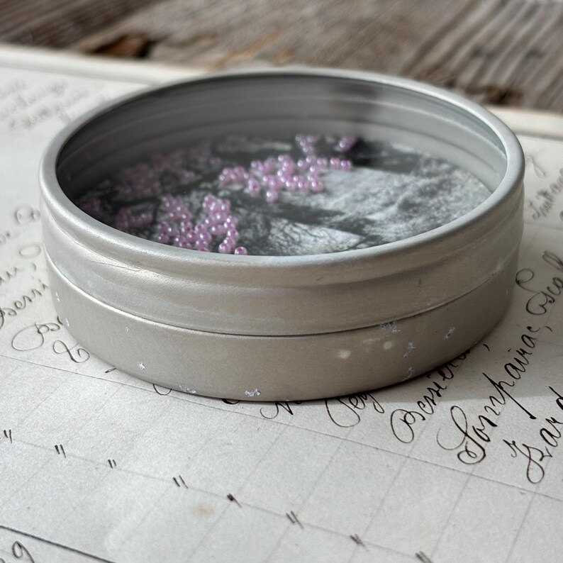 May include: A small, round, silver-colored tin with a clear lid. Inside, a grayscale image is visible, with small, light purple beads scattered across the surface. The tin sits on a sheet of paper with handwritten script.