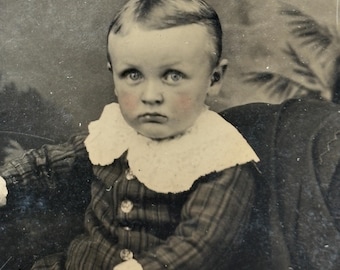 May include: Vintage black and white portrait of a young child. The child has fair skin, blue eyes, and rosy cheeks. They are wearing a plaid patterned suit with a white lace collar. The portrait is a close-up shot.