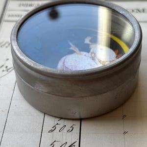 K&ouml;nnte beinhalten: Ein kleiner, runder, antiker Metallbeh&auml;lter mit einem durchsichtigen, gew&ouml;lbten Deckel. Der Deckel zeigt ein Miniaturgem&auml;lde einer Figur in einem Kleid. Der Beh&auml;lter steht auf einem Vintage-Register mit handschriftlichen Zahlen.