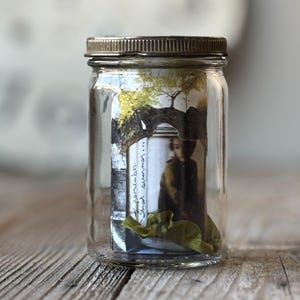 May include: A clear glass jar with a metal lid, containing a small photograph and a green ribbon. The photograph depicts a person under a tree. Handwritten text on the photograph reads "I remember...shoot summer..."