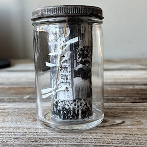 Könnte beinhalten: Ein klares Glas mit einem dunklen Metalldeckel. Im Inneren ist ein aufgerolltes Schwarz-Weiß-Bild mit japanischem Text zu sehen, zusammen mit einem dekorativen weißen und goldenen Element. Das Glas steht auf einer Holzoberfläche.