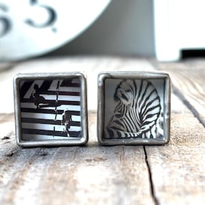 Könnte beinhalten: Zwei quadratische Manschettenknöpfe mit silberfarbenen Metallrahmen. Ein Manschettenknopf zeigt einen schwarz-weißen Zebraschädel, der andere ein schwarz-weißes Streifendesign. Die Manschettenknöpfe liegen auf einer Holzoberfläche.