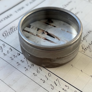 May include: A small, circular, brushed metal container with a ridged exterior. The interior holds a faded image. The container sits on a ledger with handwritten numbers and script.