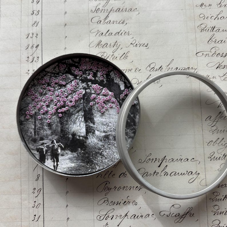 May include: A round, silver-toned container with a black and white image of a path with people walking under a tree with pink flowers. The container is open, with a clear, circular magnifying glass next to it. The background is a handwritten document.