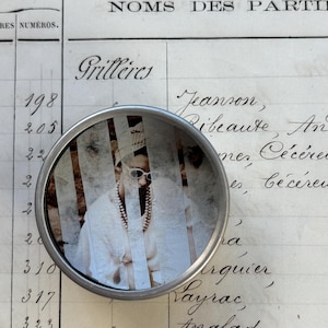 May include: A round, silver-colored magnifying glass with a vintage photograph inside. The photograph shows a person wearing sunglasses and a pearl necklace. The background is an old ledger with handwritten text.