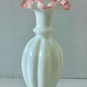 Può includere: Un vaso di vetro bianco con bordo rosa arricciato. Il vaso ha un corpo bulboso con scanalature verticali e una base svasata. Il bordo superiore del vaso ha un bordo delicato e smerlato di colore rosa tenue.