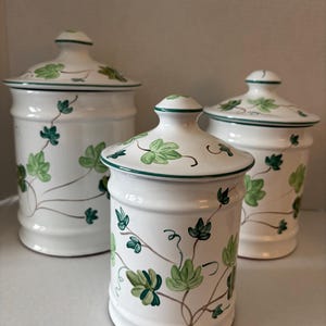 May include: Three white ceramic canisters with lids, adorned with green ivy leaf designs and green trim. The canisters vary in size and are suitable for kitchen storage.
