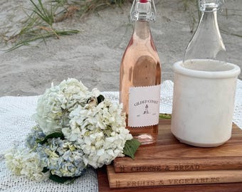 May include: A glass bottle of rose wine with a Gilded Coast lighthouse label, a clear glass bottle in a white marble cooler, and three stacked wooden cutting boards labeled Cheese & Bread, Fruits & Vegetables, and Meat & Fish, with white hydrangeas.